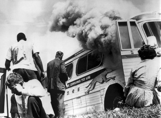 Freedom Riders sit on the ground outside the burning hulk of a Greyhound bus