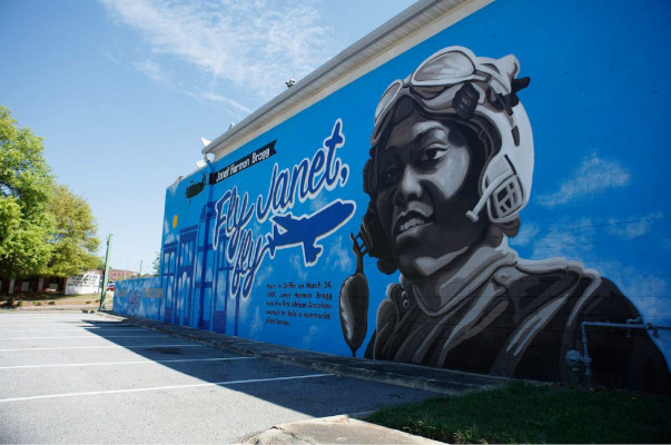 Janet Harmon Bragg mural painted on a building that sits in front of a parking lot near a post office. It shows a headshot of Janet wearing her aviation helmet and goggles against a blue sky background. A black airplane totes a banner that says Janet Harmon Bragg. A blue airplane has written "Fly Janet, fly" in the sky. Writing below the blue airplane reads: Born in Griffin on March 24, 1907, Janet Harmon Bragg was the first African American woman to hold a commercial pilot's license.