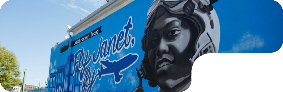 Janet Harmon Bragg mural. It shows a headshot of Janet wearing her aviation helmet and goggles against a blue sky background. A black airplane totes a banner that says Janet Harmon Bragg. A blue airplane has written "Fly Janet, fly" in the sky.