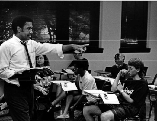 James E. McLeod holding a book and pointing in a classroom as students view textbooks on their desks.